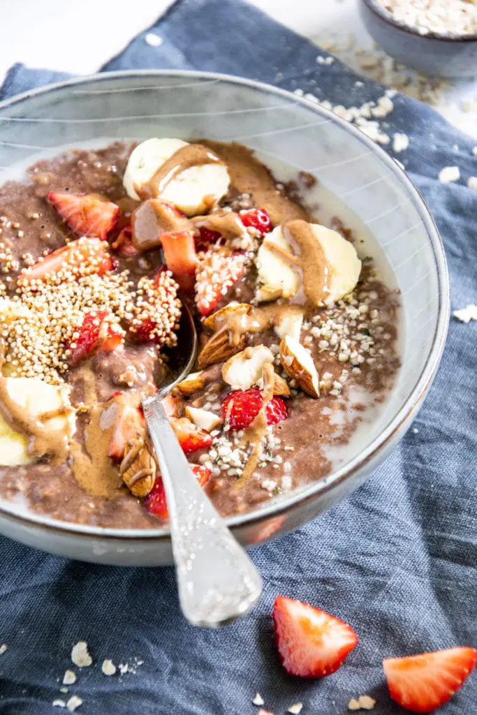 Schoko-Mandel-Porridge getoppt mit Bananenscheiben, Erdbeeren, Mandeln und Mandelmus. In einer blaugrauen Schüssel mit Löffel darin. 45-Grad-Winkel