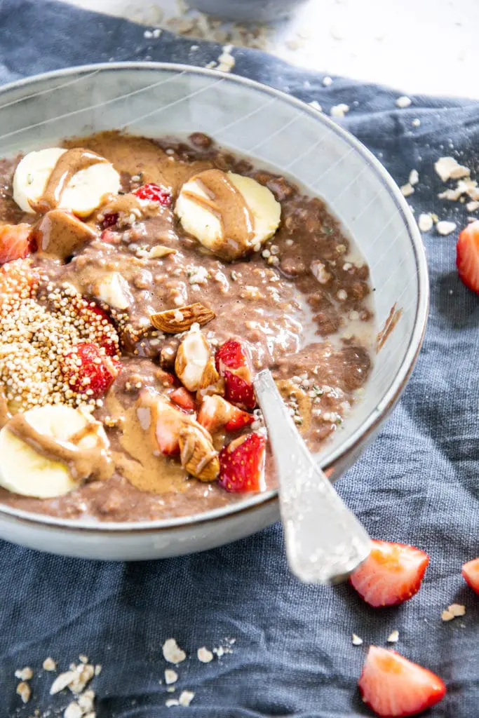 Schoko-Mandel-Porridge getoppt mit Bananenscheiben, Erdbeeren, Mandeln und Mandelmus. In einer blaugrauen Schüssel mit Löffel darin. 45-Grad-Winkel.