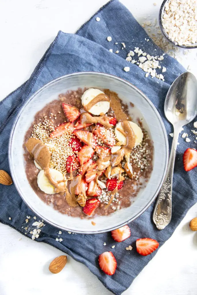 Cremiger Schoko-Mandel-Porridge getoppt mit Bananenscheiben, Erdbeeren, Mandeln und Mandelmus. In einer blaugrauen Schüssel mit Löffel darin, auf einem blauen Leinentuch. Top View.