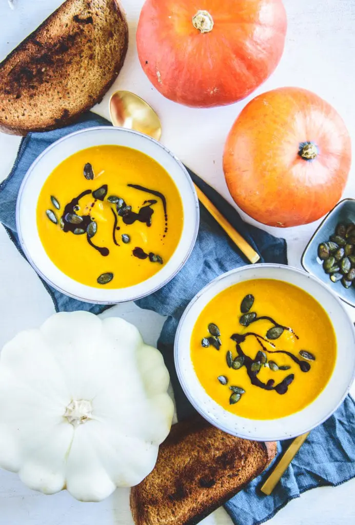 Zwei helle, grau-blaue Schüsseln mit tiefgelber Kürbissuppe, garniert mit Kürbiskernen und Kürbiskernöl. Von oben fotografiert mit einem blauen Leinentuch und Kürbisse als Dekoration.