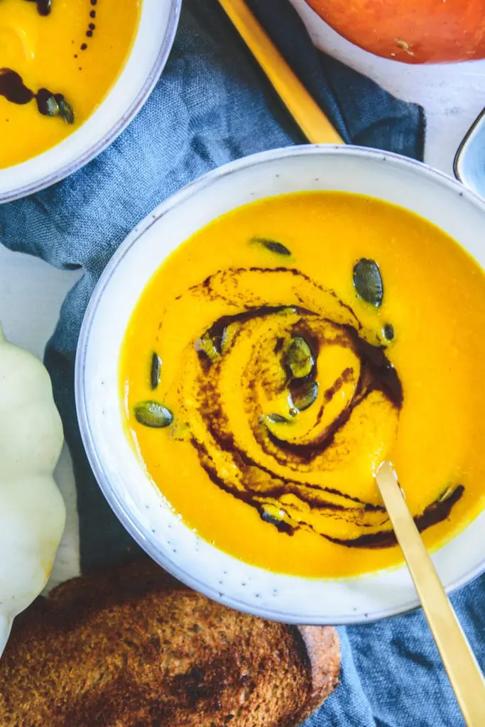 Sehr gelb-orange, cremige Kürbissuppe mit Kernöl marmoriert und mit Kürbiskernen bestreut in einer hellen, grau-blauen Schüssel mit einem goldenen Löffel darin.