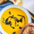 Sehr gelb-orange, cremige Kürbissuppe mit Kernöl marmoriert und mit Kürbiskernen bestreut in einer hellen, grau-blauen Schüssel mit einer Scheibe geröstetem Brot am Rand