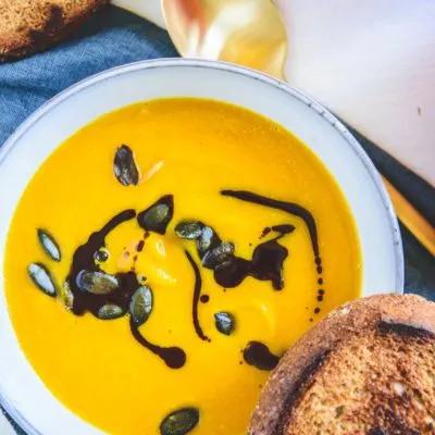 Sehr gelb-orange, cremige Kürbissuppe mit Kernöl marmoriert und mit Kürbiskernen bestreut in einer hellen, grau-blauen Schüssel mit einer Scheibe geröstetem Brot am Rand