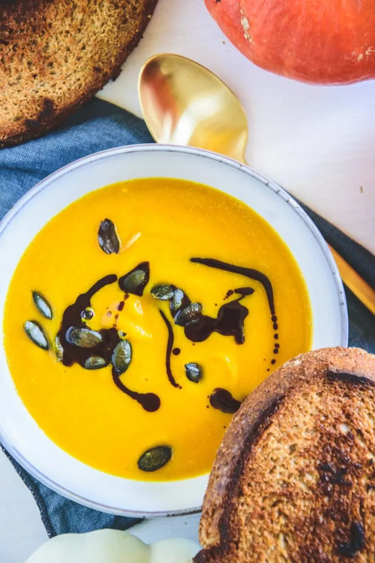 Sehr gelb-orange, cremige Kürbissuppe mit Kernöl marmoriert und mit Kürbiskernen bestreut in einer hellen, grau-blauen Schüssel mit einer Scheibe geröstetem Brot am Rand