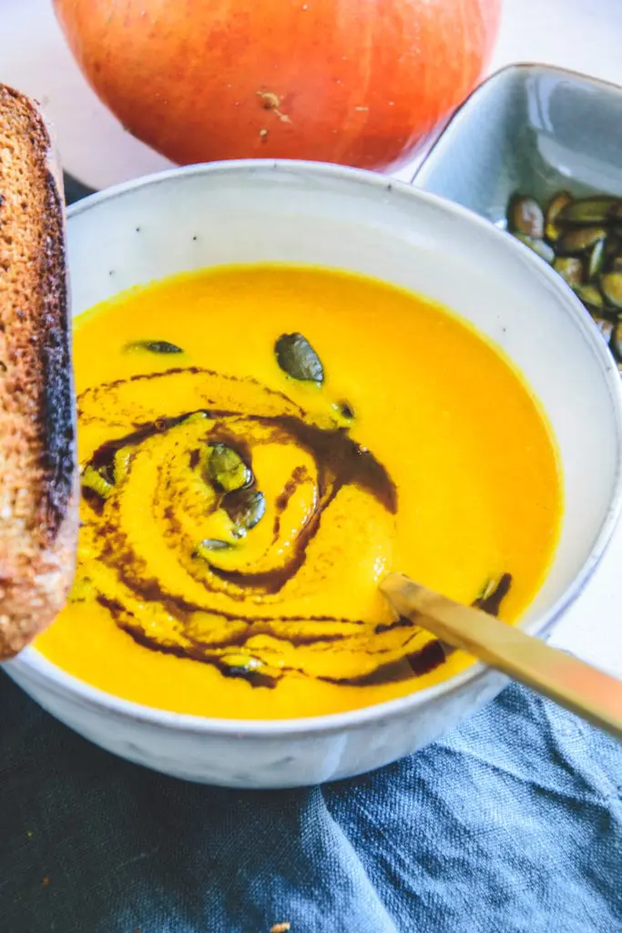 Sehr gelb-orange, cremige Kürbissuppe mit Kernöl marmoriert und mit Kürbiskernen bestreut in einer hellen, grau-blauen Schüssel mit einem goldenen Löffel darin und einer Scheibe geröstetem Brot am Rand