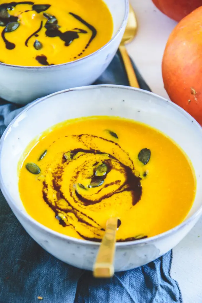 Sehr gelb-orange, cremige Kürbissuppe mit Kernöl marmoriert und mit Kürbiskernen bestreut in einer hellen, grau-blauen Schüssel.