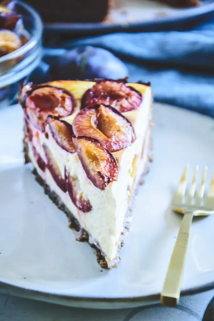 Ein Stück gesunder Zwetschgenkuchen mit Topfen und Nüssen im 45-Grad-Winkel auf einem grau-blauen, hellen Teller mit einer goldenen Gabel daneben.