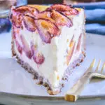 Ein Stück gesunder Zwetschgenkuchen mit Topfen und Nüssen im 45-Grad-Winkel auf einem grau-blauen, hellen Teller mit einer goldenen Gabel daneben.
