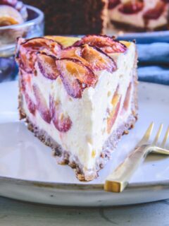 Ein Stück gesunder Zwetschgenkuchen mit Topfen und Nüssen im 45-Grad-Winkel auf einem grau-blauen, hellen Teller mit einer goldenen Gabel daneben.