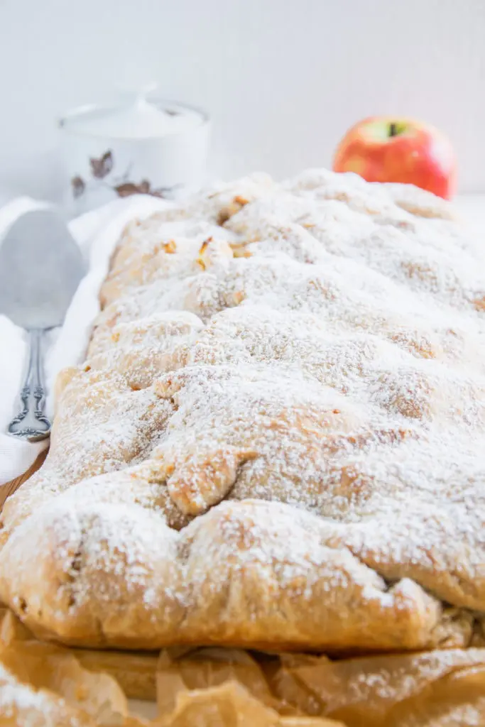 Gesunde Apfelschlangerl mit Puder-Erythrit bestreut auf braunem Backpapier und einem großen Holzbrett. Weißes Tuch, Tortenheber, Zuckerdose und Apfel im Hintergrund. Aufnahme im 45-Grad-Winkel.