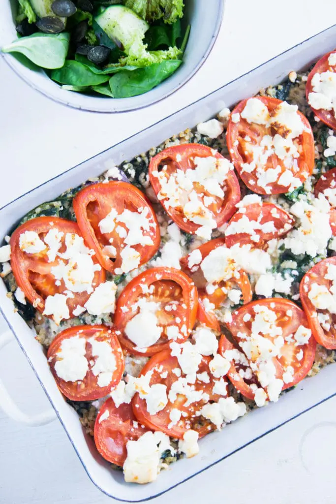 Gesunder Bulgur-Spinatauflauf mit Kichererbsen, Tomaten und Feta in einer weißen Emaille-Auflaufform. Aufnahme von oben auf weißem Untergrund. Schüssel mit Salat neben der Auflaufform.