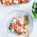 Ein Stück Gesunder Bulgur-Spinatauflauf mit Kichererbsen, Tomaten und Feta auf einem graublauen Teller mit einer goldenen Gabel und einer Salatschüssel daneben auf weißem Untergrund. Aufnahme im Top View.