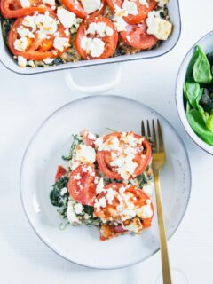 Ein Stück Gesunder Bulgur-Spinatauflauf mit Kichererbsen, Tomaten und Feta auf einem graublauen Teller mit einer goldenen Gabel und einer Salatschüssel daneben auf weißem Untergrund. Aufnahme im Top View.