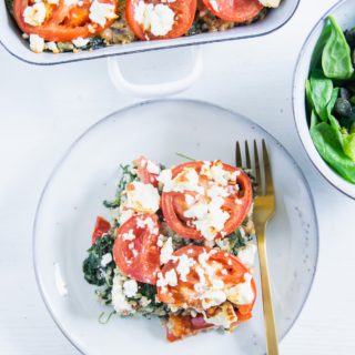 Ein Stück Gesunder Bulgur-Spinatauflauf mit Kichererbsen, Tomaten und Feta auf einem graublauen Teller mit einer goldenen Gabel und einer Salatschüssel daneben auf weißem Untergrund. Aufnahme im Top View.