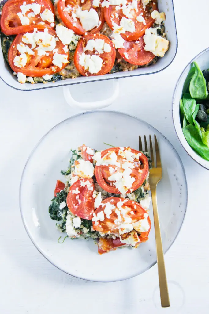 Ein Stück Gesunder Bulgur-Spinatauflauf mit Kichererbsen, Tomaten und Feta auf einem graublauen Teller mit einer goldenen Gabel und einer Salatschüssel daneben auf weißem Untergrund. Aufnahme im Top View.