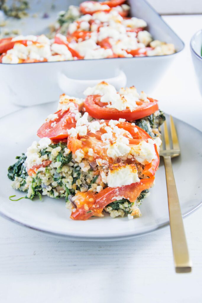 Ein Stück Gesunder Bulgur-Spinatauflauf mit Kichererbsen, Tomaten und Feta auf einem graublauen Teller mit einer goldenen Gabel daneben. Aufnahme im 45-Grad-Winkel.