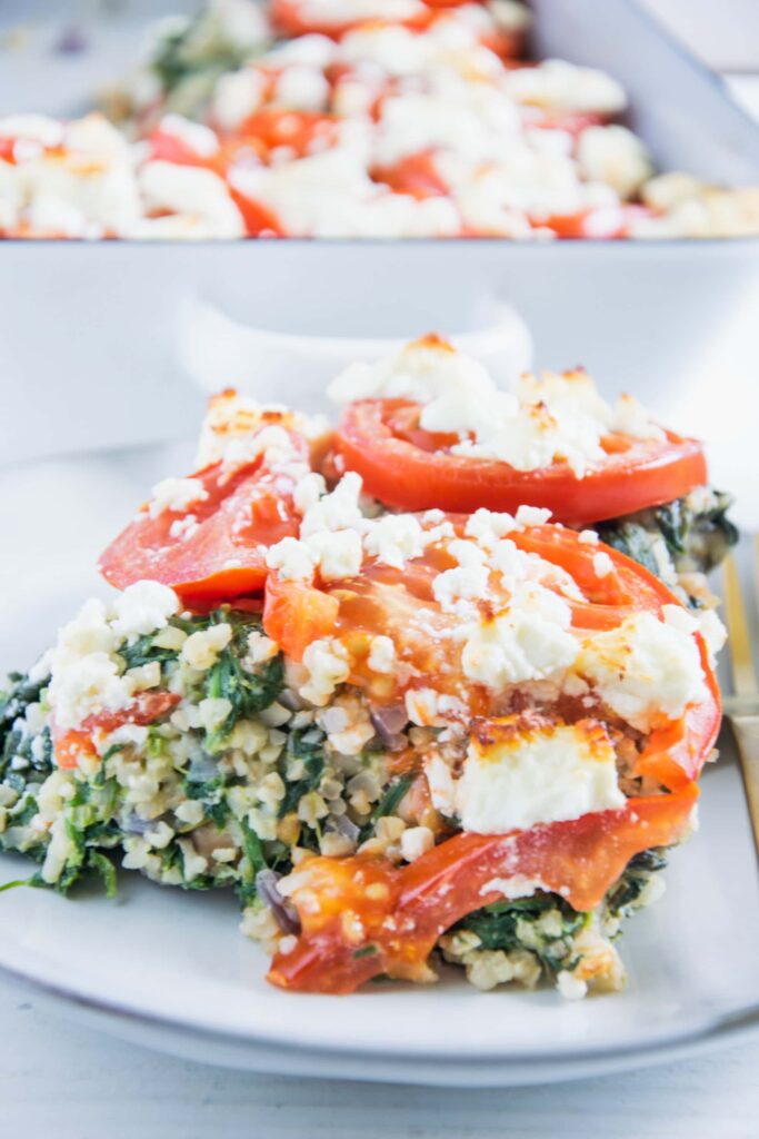 Ein Stück gesunder Bulgur-Spinatauflauf mit Kichererbsen, Tomaten und Feta auf einem graublauen Teller mit einer goldenen Gabel daneben. Aufnahme im 45-Grad-Winkel.