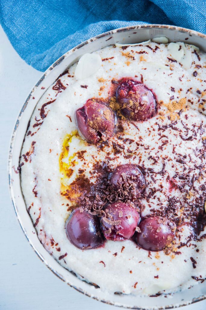 Dinkel-Grießbrei mit Schokolade und Kirschen in einer grauen Schüssel mit einem goldenen Löffel. Aufnahme von oben, angeschnitten von rechts.