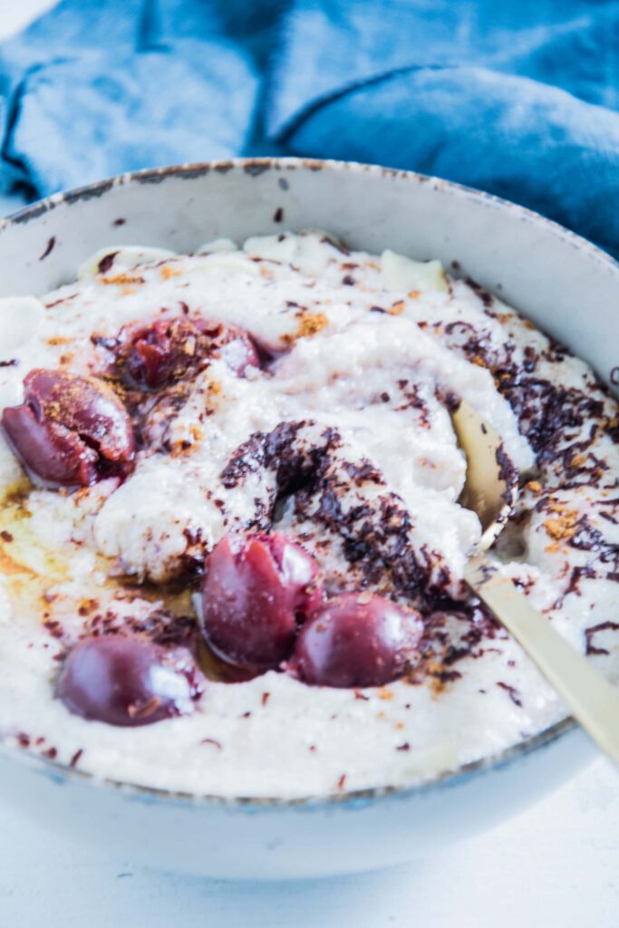 Dinkel-Grießbrei mit Schokolade und Kirschen in einer grauen Schüssel mit einem goldenen Löffel. Nahaufnahme im 45-Grad-Winkel mit vielen Details. Der Löffel hat den Brei leicht vermischt.