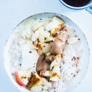 Klassischer Porridge aus Haferflocken und Milch mit Apfel, Mandelmus, Mandeblättchen, Joghurt, Samen und Zimt als Topping. In einer hellen Schüssel mit Löffel darin. Aufnahme im Top View.