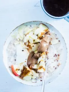 Klassischer Porridge aus Haferflocken und Milch mit Apfel, Mandelmus, Mandeblättchen, Joghurt, Samen und Zimt als Topping. In einer hellen Schüssel mit Löffel darin. Aufnahme im Top View.