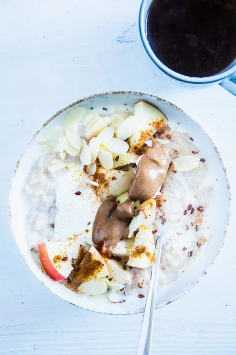 Klassischer Porridge aus Haferflocken und Milch mit Apfel, Mandelmus, Mandeblättchen, Joghurt, Samen und Zimt als Topping. In einer hellen Schüssel mit Löffel darin. Aufnahme im Top View.