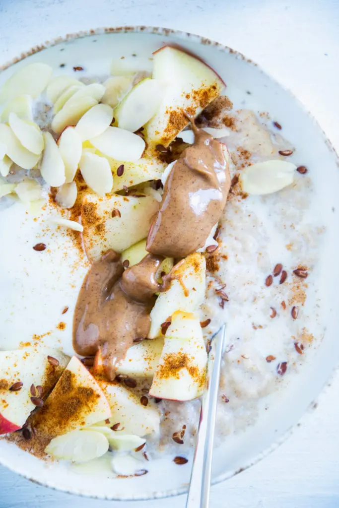 Klassischer Porridge aus Haferflocken und Milch mit Apfel, Mandelmus, Mandeblättchen, Joghurt, Samen und Zimt als Topping. In einer hellen Schüssel mit Löffel darin. Nahaufnahme im Top View.