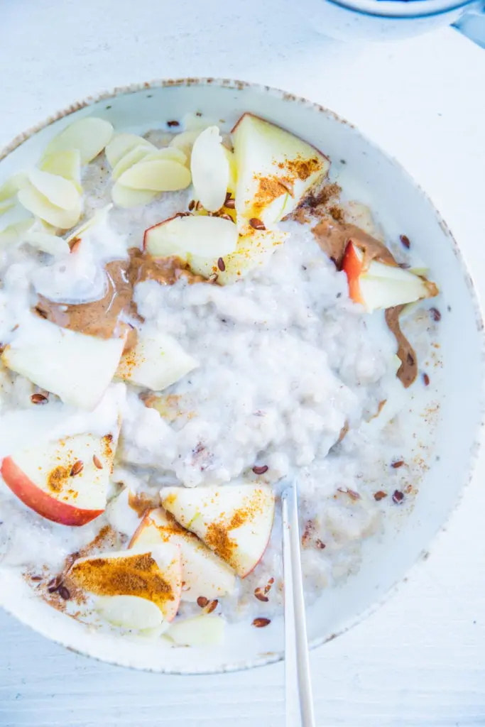 Klassischer Porridge aus Haferflocken und Milch mit Apfel, Mandelmus, Mandeblättchen, Joghurt, Samen und Zimt als Topping. In einer hellen Schüssel mit Löffel darin. Nahaufnahme im Top View.