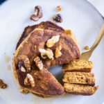 Gestapelte Kürbis-Pancakes von oben auf einem graublauen Teller. Gehackte Walnüsse als Topping. Eine goldene Gabel hat ein herausgeschnittenes Tortenstück aus dem Stapel aufgespießt und liegt neben dem Stapel.