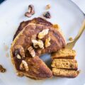 Gestapelte Kürbis-Pancakes von oben auf einem graublauen Teller. Gehackte Walnüsse als Topping. Eine goldene Gabel hat ein herausgeschnittenes Tortenstück aus dem Stapel aufgespießt und liegt neben dem Stapel.