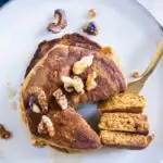 Gestapelte Kürbis-Pancakes von oben auf einem graublauen Teller. Gehackte Walnüsse als Topping. Eine goldene Gabel hat ein herausgeschnittenes Tortenstück aus dem Stapel aufgespießt und liegt neben dem Stapel.