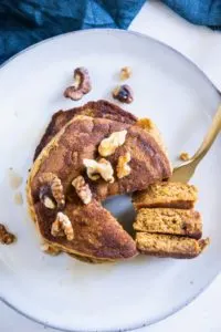 Gestapelte Kürbis-Pancakes von oben auf einem graublauen Teller. Gehackte Walnüsse als Topping. Eine goldene Gabel hat ein herausgeschnittenes Tortenstück aus dem Stapel aufgespießt und liegt neben dem Stapel.