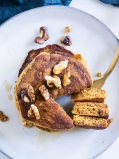 Gestapelte Kürbis-Pancakes von oben auf einem graublauen Teller. Gehackte Walnüsse als Topping. Eine goldene Gabel hat ein herausgeschnittenes Tortenstück aus dem Stapel aufgespießt und liegt neben dem Stapel.