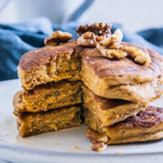 Drei vegane Kürbis-Pancakes gestapelt und angeschnitten mit Walnüssen und Ahornsirup. Aufnahme auf Augenhöhe.