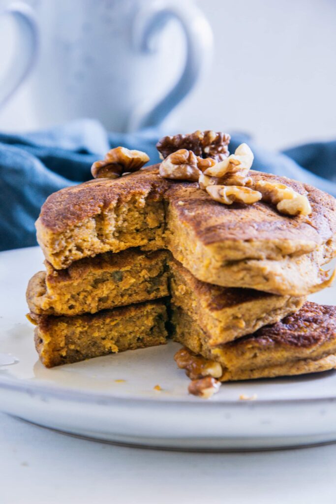 Drei vegane Kürbis-Pancakes gestapelt und angeschnitten mit Walnüssen und Ahornsirup. Aufnahme auf Augenhöhe.