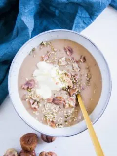 Maroni-Kartoffelsuppe in einer graublauen Schüssel mit einem goldenen Löffel. Garniert mit Maronistücken, Hanfsamen und Crème fraîche. Aufnahme von oben.