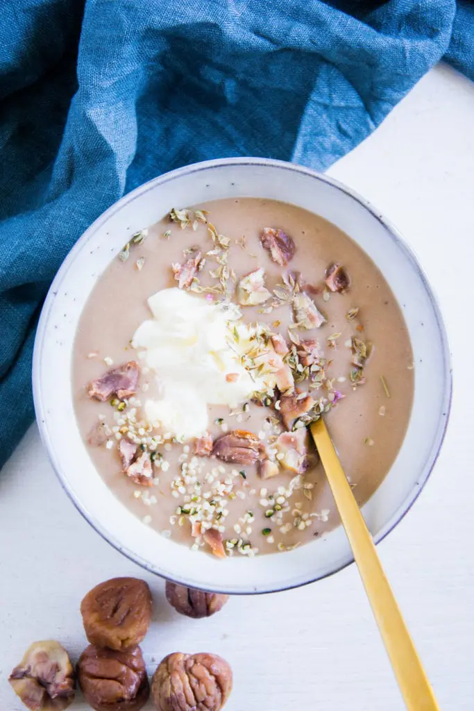 Maroni-Kartoffelsuppe in einer graublauen Schüssel mit einem goldenen Löffel. Garniert mit Maronistücken, Hanfsamen und Crème fraîche. Aufnahme von oben.