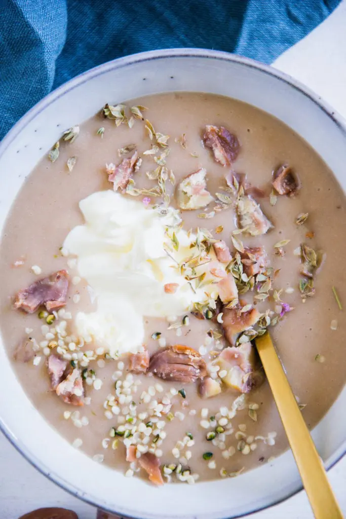 Maroni-Kartoffelsuppe in einer graublauen Schüssel mit einem goldenen Löffel. Garniert mit Maronistücken, Hanfsamen und Crème fraîche. Nahaufnahme von oben.