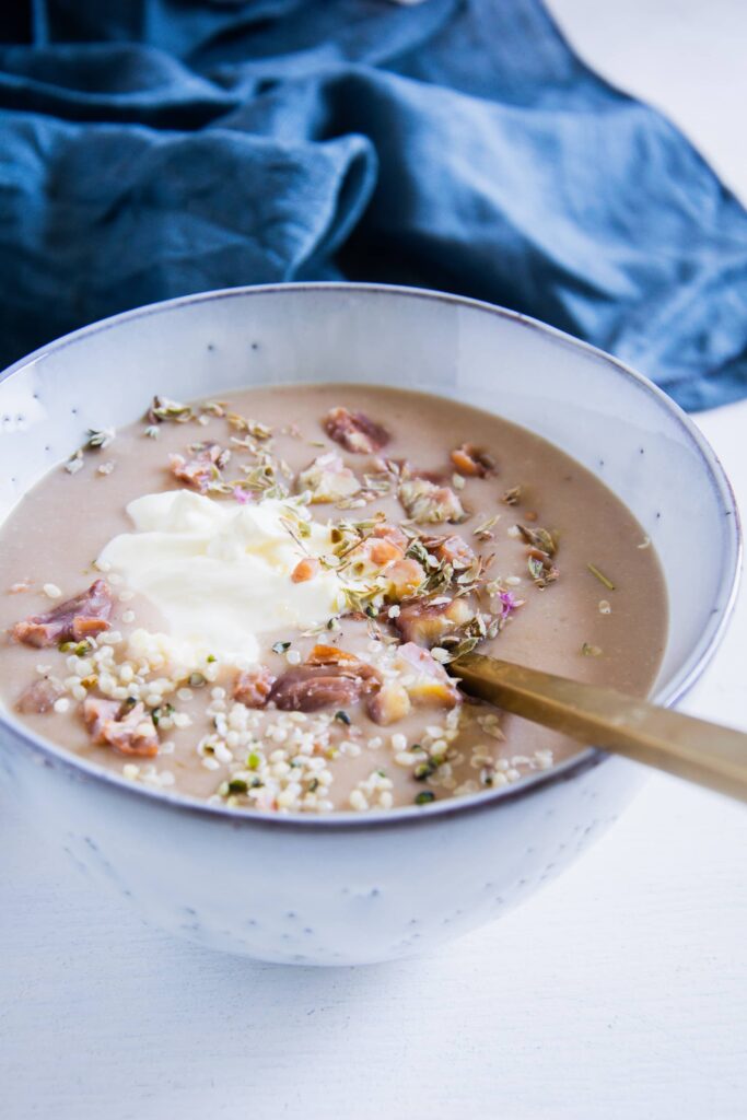 Maroni-Kartoffelsuppe in einer graublauen Schüssel mit einem goldenen Löffel. Garniert mit Maronistücken, Hanfsamen und Crème fraîche. Aufnahme im 45-Grad-Winkel