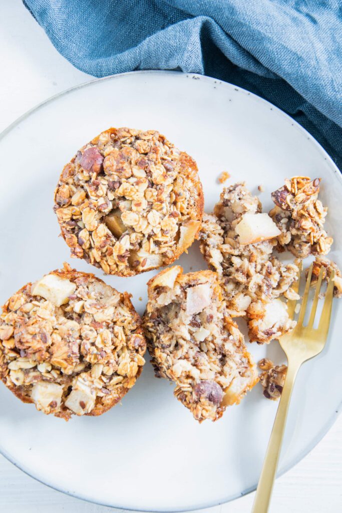 Drei Oatmeal Muffins auf einem blaugrauen Teller mit einer goldenen Gabel, die einen von ihnen in der Mitte zerteilt hat, sodass das Innere sichtbar ist.