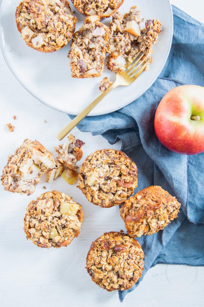 Oatmeal Muffins mit Apfel und Nüssen von oben auf weißem Untergrund mit blauem Leinentuch und einem Apfel. Die Oatmeal Muffins sind von oben sichtbar oder liegen auf der Seite. Einer ist auseinander gebrochen.