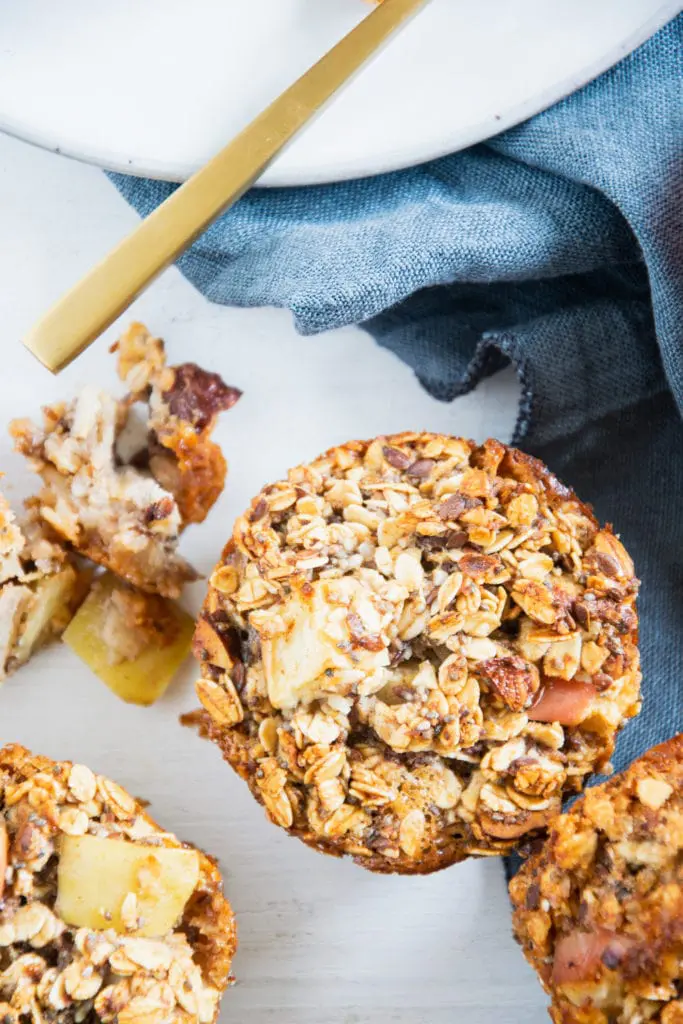Ein Oatmeal Muffin mit Apfel und Nüssen in der Nahaufnahme von oben auf weißem Untergrund mit blauem Leinentuch. Stücke eines zweiten Muffins liegen daneben.