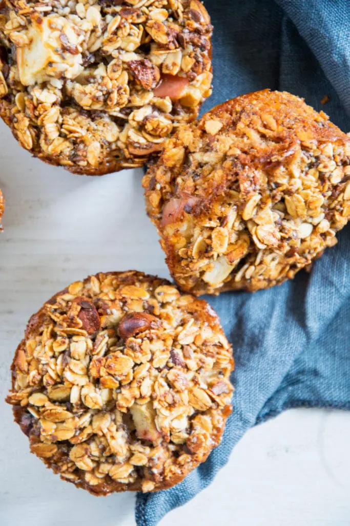 Oatmeal Muffins mit Apfel und Nüssen in der Nahaufnahme von oben auf weißem Untergrund mit blauem Leinentuch. Von zwei Muffins ist die Oberfläche sichtbar, der dritte liegt auf der Seite.