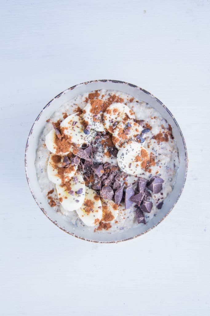 Porridge getoppt mit Bananenscheiben, gehackter Schokolade, Kakaopulver, Kakaonibs und Chiasamen. Aufnahme von oben.