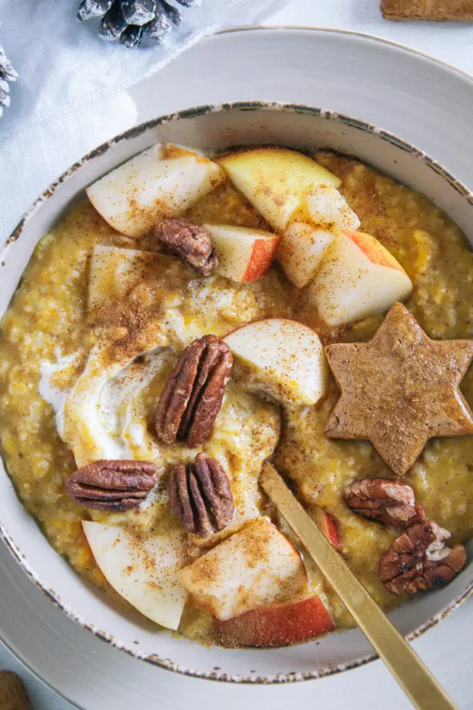 Kürbis-Porridge mit einem Topping aus Apfelstücken, Pekannüssen, einem Lebkuchenstern und Zimt. Goldener Apfel im Porridge. Nahaufnahme von oben