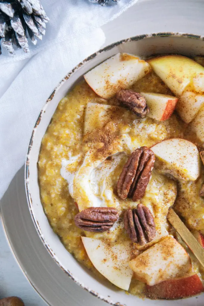 Kürbis-Porridge mit einem Topping aus Apfelstücken, Pekannüssen, einem Lebkuchenstern und Zimt. Goldener Apfel im Porridge. Nahaufnahme von oben, rechts angeschnitten.