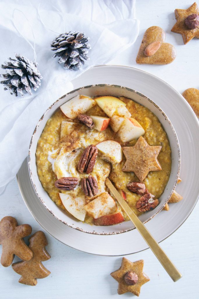 Kürbis-Porridge mit einem Topping aus Apfelstücken, Pekannüssen, einem Lebkuchenstern und Zimt. Goldener Apfel im Porridge. Braune Schüssel auf beigem Teller und weißem Untergrund. Dekoriert mit einem weißen Leinentuch und Zapfen.