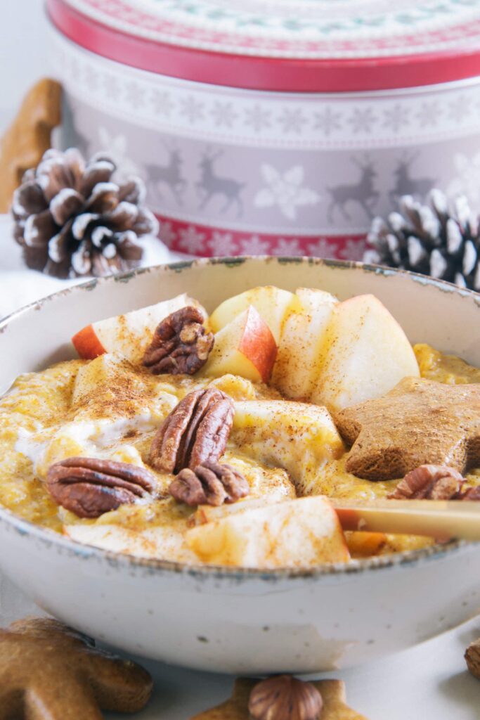 Kürbis-Porridge mit einem Topping aus Apfelstücken, Pekannüssen, einem Lebkuchenstern und Zimt. Goldener Apfel im Porridge. Aufnahme im 45-Grad-Winkel mit einer Keksdose und Zapfen im Hintergrund.