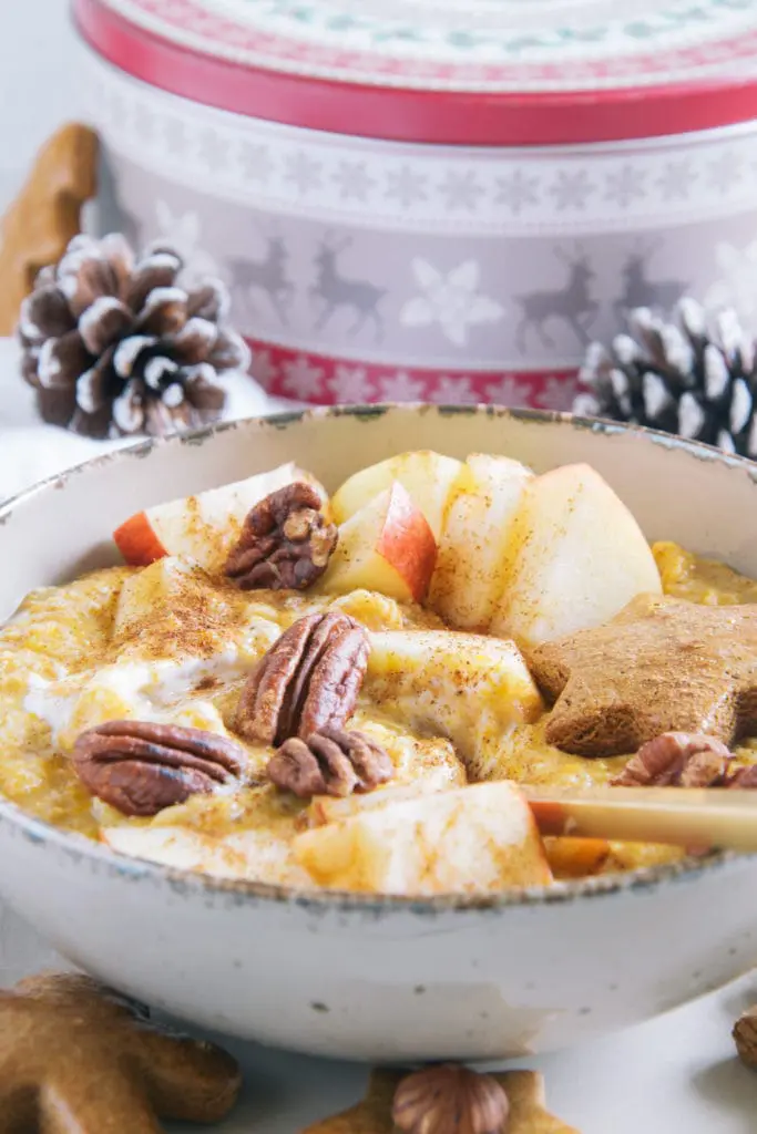 Kürbis-Porridge mit einem Topping aus Apfelstücken, Pekannüssen, einem Lebkuchenstern und Zimt. Goldener Apfel im Porridge. Aufnahme im 45-Grad-Winkel mit einer Keksdose und Zapfen im Hintergrund.