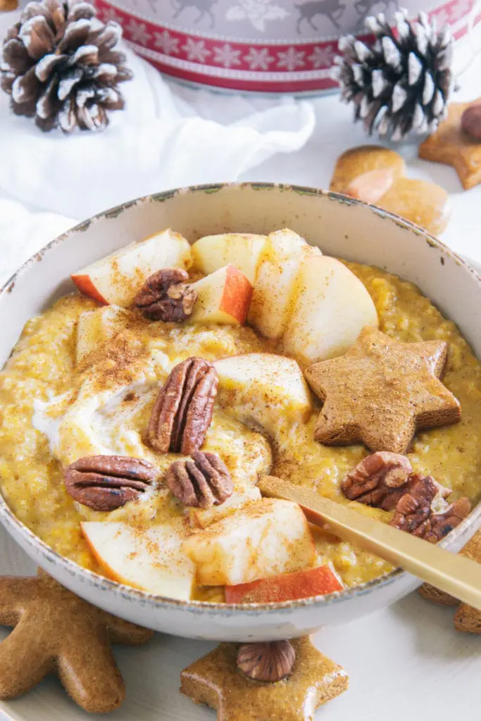 Kürbis-Porridge mit einem Topping aus Apfelstücken, Pekannüssen, einem Lebkuchenstern und Zimt. Goldener Apfel im Porridge. Aufnahme im 45-Grad-Winkel.
